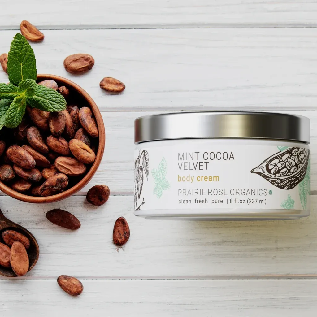 Photorealistic 3D packaging rendering for Prairie Rose Organics Mint Cocoa Velvet body cream jar on a white wood background with cocoa beans.