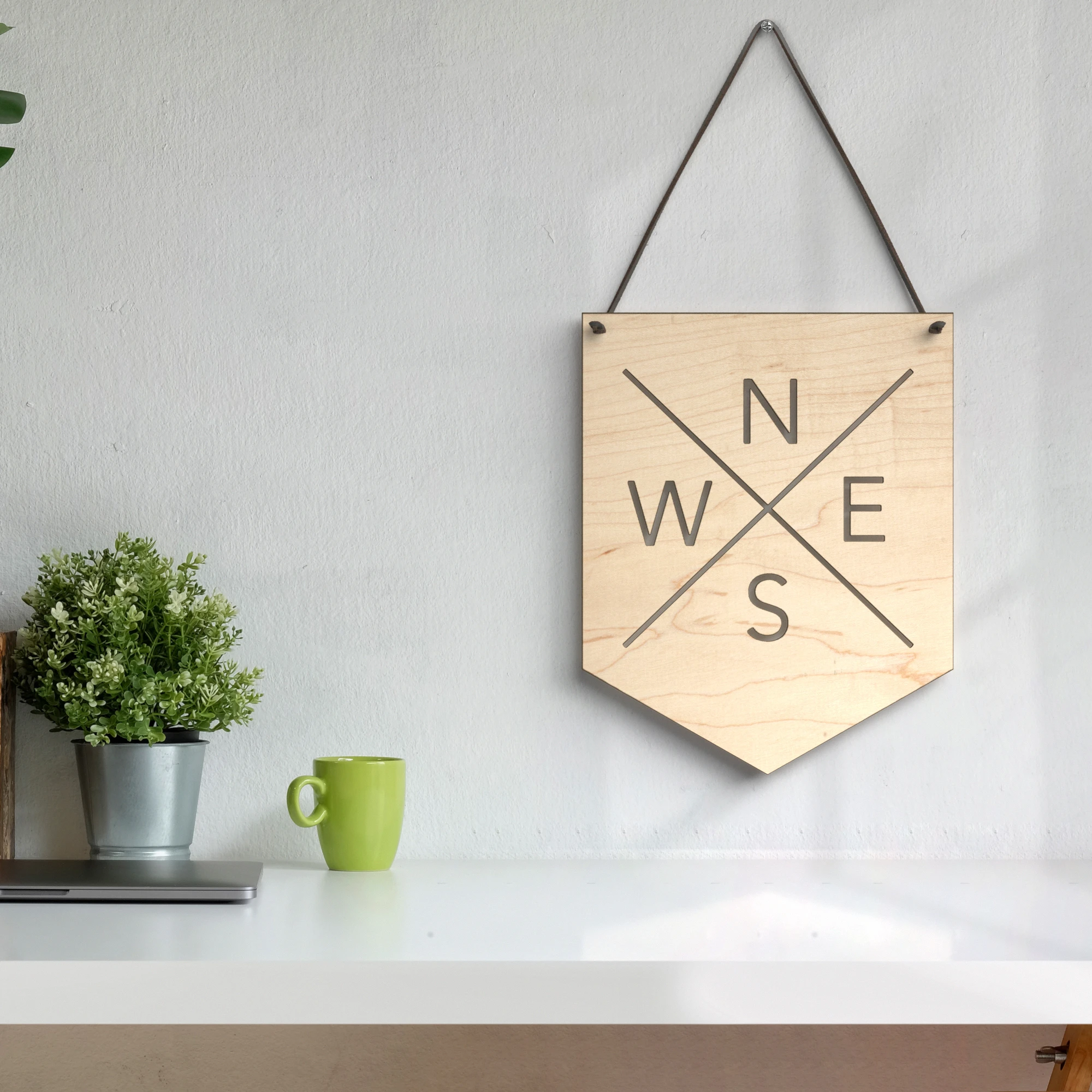 3D lifestyle rendering of a wooden compass wall board hanging above a clean white desk with a laptop, green mug, and small potted plant.