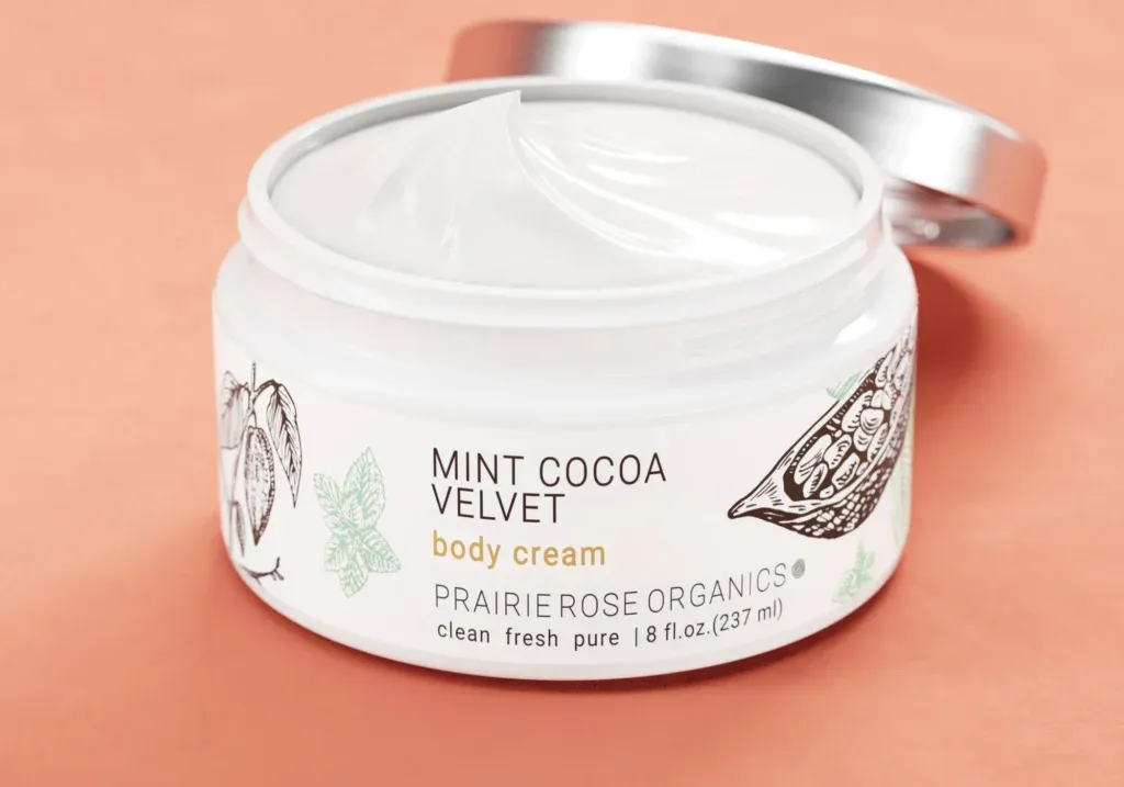 Close-up packaging 3d rendering of Prairie Rose Organics Mint Cocoa Velvet body cream jar with an open lid and realistic cream texture.