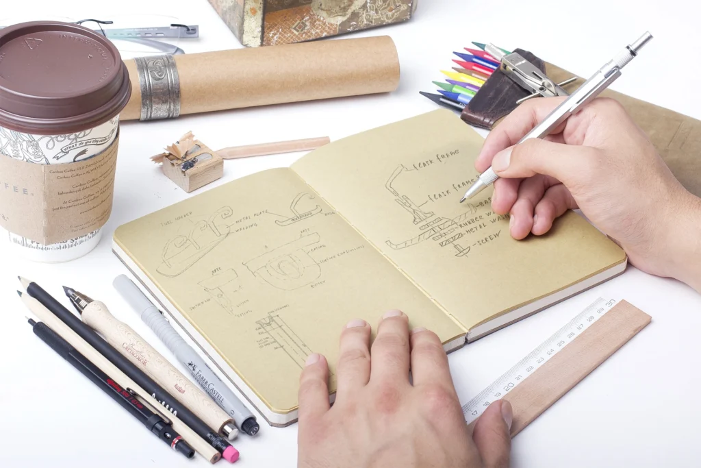 A top-down view of a designer sketching technical product diagrams for a hummingbird feeder in a brown paper notebook, surrounded by drawing tools, a wooden ruler, and a coffee cup.