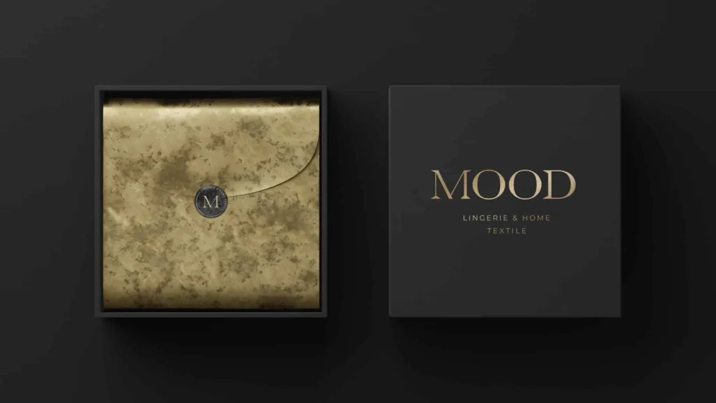 Top-down view of a luxury matte black gift box packaging mockup for Mood Lingerie & Home Textile, showing an open box with gold foil wrapping paper secured by an M monogram sticker next to the closed box lid featuring a gold serif logo.