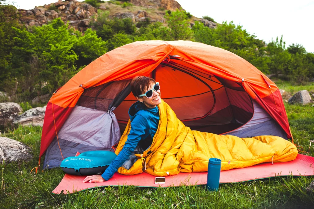 Smiling woman in a sleeping bag outside an orange tent using the Pitch and Trek camping pillow at a scenic outdoor campsite